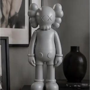 May include: A gray vinyl figure with a skull-like face and crossed-out eyes. The figure has rounded ears and a simple, stylized body. It stands on a black surface, next to a black vase and a framed picture.