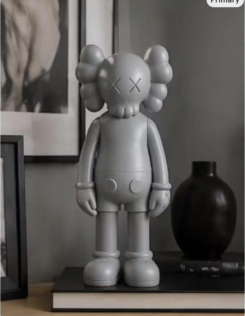 May include: A gray vinyl figure with a skull-like face and crossed-out eyes. The figure has rounded ears and a simple, stylized body. It stands on a black surface, next to a black vase and a framed picture.