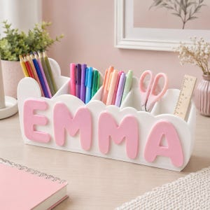 Puede incluir: Un organizador de escritorio blanco con bordes festoneados y el nombre EMMA en letras rosas grandes y en relieve en la parte frontal. El organizador tiene varios compartimentos con lápices de colores, rotuladores, tijeras rosas y una regla de madera.