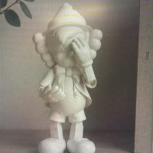 K&ouml;nnte beinhalten: Eine wei&szlig;e Figur einer Zeichentrickfigur mit durchgestrichenen Augen, die ihr Gesicht mit einer Hand bedeckt. Die Figur tr&auml;gt einen Hut, Shorts und eine Jacke. Die Figur steht auf einem Regal.
