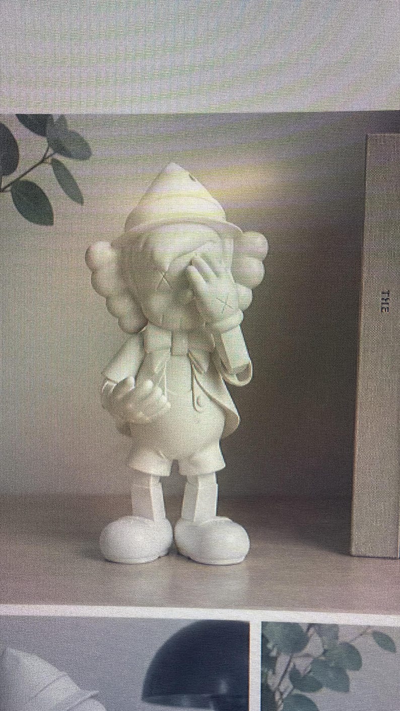 K&ouml;nnte beinhalten: Eine wei&szlig;e Figur einer Zeichentrickfigur mit durchgestrichenen Augen, die ihr Gesicht mit einer Hand bedeckt. Die Figur tr&auml;gt einen Hut, Shorts und eine Jacke. Die Figur steht auf einem Regal.