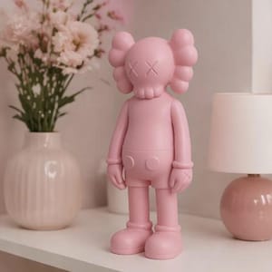 May include: A solid pink figurine with a skull-like face and crossed-out eyes stands on a white shelf. The figurine has rounded features and is wearing shorts. A vase of pink flowers and a pink lamp are in the background.