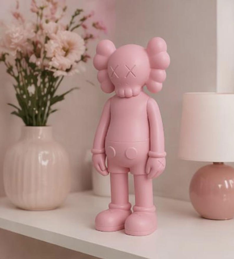 May include: A solid pink figurine with a skull-like face and crossed-out eyes stands on a white shelf. The figurine has rounded features and is wearing shorts. A vase of pink flowers and a pink lamp are in the background.