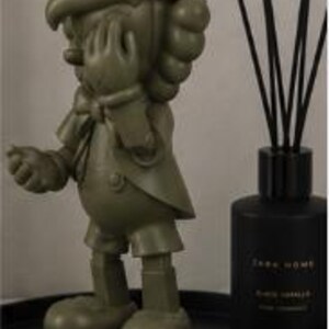 K&ouml;nnte beinhalten: Eine olivgr&uuml;ne Pinocchio-Figur steht auf einem schwarzen Tablett neben einem schwarzen Zara Home Diffusor. Die Figur befindet sich in einer klassischen Pose, mit einer Hand zum Gesicht erhoben. Der Diffusor hat schwarze St&auml;bchen.