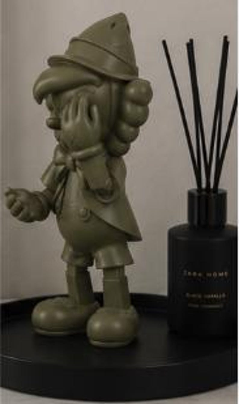 K&ouml;nnte beinhalten: Eine olivgr&uuml;ne Pinocchio-Figur steht auf einem schwarzen Tablett neben einem schwarzen Zara Home Diffusor. Die Figur befindet sich in einer klassischen Pose, mit einer Hand zum Gesicht erhoben. Der Diffusor hat schwarze St&auml;bchen.