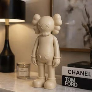 May include: A beige vinyl figurine with crossed-out eyes and a rounded body. The figurine is standing on a white surface, with a black lamp and books with the titles "CHANEL" and "TOM FORD" in the background.