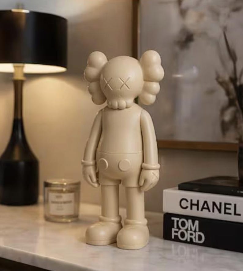 May include: A beige vinyl figurine with crossed-out eyes and a rounded body. The figurine is standing on a white surface, with a black lamp and books with the titles "CHANEL" and "TOM FORD" in the background.