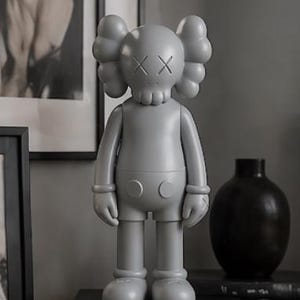 May include: A grey vinyl figure with a skull-like face and crossed-out eyes. The figure has rounded ears and a stylized body. It stands on a black surface, next to a black vase and framed artwork. A modern art piece.