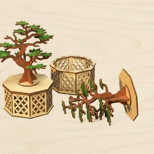May include: Wooden bonsai tree model with a brown trunk and green leaves, displayed on a laser-cut wooden base. The image also shows a separate wooden container and a tree component.
