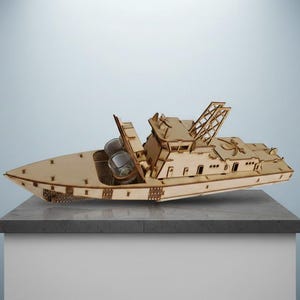May include: A wooden model of a boat with a detailed design, including a cabin, mast, and open deck. The boat is made of light-colored wood and sits on a gray surface. Two clear glasses are visible on the deck.