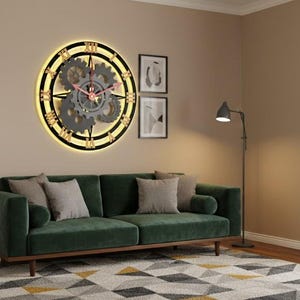 May include: A large, round wall clock with a gear design and Roman numerals. The clock is illuminated with a warm light and hangs above a green velvet sofa. The room also features a side table with a plant and a floor lamp.
