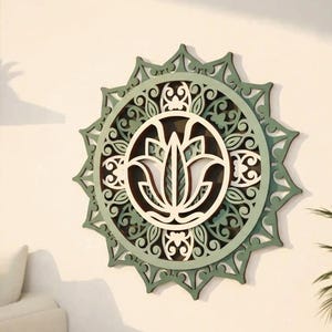 May include: A circular wall art piece featuring a layered, laser-cut design. The centre displays a stylised lotus flower in cream, surrounded by intricate, symmetrical scrollwork patterns in shades of sage green and dark brown, all set within a star-shaped frame.