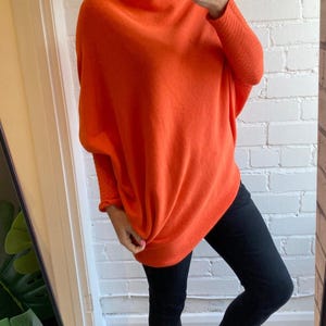 Könnte beinhalten: Ein leuchtend orangefarbener, übergroßer Pullover mit weitem Ausschnitt und langen Ärmeln. Die Ärmel haben ein strukturiertes Strickmuster. Kombiniert mit einer schwarzen Hose und Sandalen. Ein auffälliges Outfit.