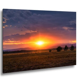 Könnte beinhalten: Ein lebendiger Sonnenuntergang über einem Feld und Bergen. Der Himmel wechselt von tiefem Blau zu feurigem Orange und Gelb, mit strukturierten Wolken. Silhouetten von Bäumen und einem Zaun säumen den Horizont und schaffen eine ruhige Landschaft.