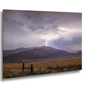 May include: A landscape photograph depicts a dramatic thunderstorm over a mountain range. Lightning strikes illuminate the dark, cloudy sky. The foreground shows a field of dry grass and a wooden fence.