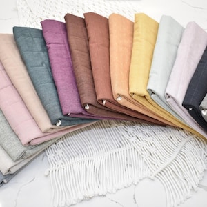 May include: A collection of folded fabric squares in various colors, including gray, pink, purple, and yellow. The fabrics are arranged in a fan shape, showcasing their different hues and textures. The arrangement is on a white surface.