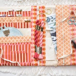 May include: A fabric craft organizer roll featuring orange, pink, and cream geometric patterns. It includes pockets holding embroidery floss, a small hoop with a cat illustration, a needle, a zipper pouch, scissors, and a black marking pen.