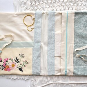 May include: Cream-colored jewelry organizer with floral embroidery and multiple compartments. Features a light blue zipper and accents, along with a patterned fabric section. Includes a drawstring closure and gold-colored ring holders.