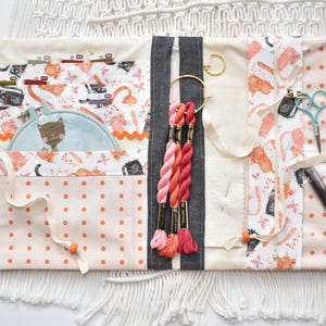 May include: A fabric craft organizer featuring a cat-patterned print, polka dot sections, and macrame fringe. It includes pockets holding embroidery floss, a small hoop with a cat illustration, a needle cushion, scissors, and a black pen.