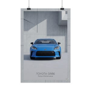 Könnte beinhalten: Ein vertikales Poster mit einem hellblauen Toyota GR86 Sportwagen in der Frontansicht. Der Text TOYOTA GR86 Purity of Performance ist unten aufgedruckt. Das Auto ist vor einer hellgrauen Betonwand mit geometrischen Schatten positioniert.