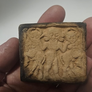 May include: A small, square, tan-colored relief carving featuring three figures. The figures are stylized with detailed facial features and appear to be in a procession or dance. The edges of the carving are dark brown.