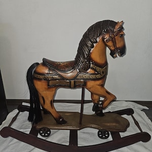 May include: A wooden rocking horse with a light brown body, dark brown mane, and a black tail. The horse features a detailed saddle with decorative patterns, a bridle, and is mounted on a platform with four small wheels attached to curved rockers.