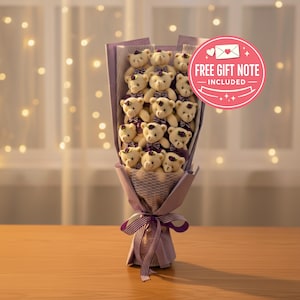 Peut inclure: Un bouquet de petits ours en peluche enveloppés dans du papier violet et blanc. Chaque ours porte un nœud papillon violet. Le bouquet est noué avec un ruban violet. Un cercle rose avec le texte "FREE GIFT NOTE INCLUDED" est visible.
