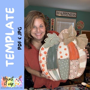 Puede incluir: Un recorte decorativo de madera con forma de calabaza que presenta un diseño de patchwork con segmentos en naranja, verde salvia y crema. La calabaza está adornada con patrones pintados de puntos, estrellas y flores, y un gran lazo de arpillera.