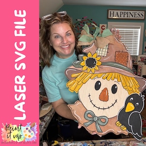 May include: A wooden scarecrow door hanger featuring a brown hat with a sunflower, yellow straw hair, a stitched nose, and a black crow. The design includes a green bow at the neck and a burlap ribbon topper. The text LASER SVG FILE is visible.