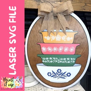 May include: Wooden sign featuring a stack of colorful casserole dishes with floral designs. The sign has a burlap bow at the top and a white border. The text "LASER SVG FILE" is on the left side.