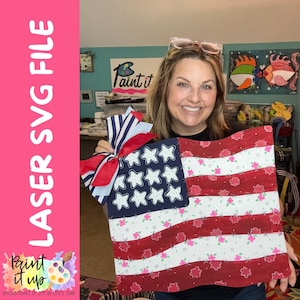 May include: A decorative wooden American flag with red and white stripes, blue star field, and a patriotic bow. The flag is held by a person. The image includes the text "LASER SVG FILE" and "Paint it up".