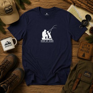 Peut inclure: T-shirt bleu marine avec un motif blanc d'une personne et d'un enfant pêchant, avec le texte "SIDE BY SIDE". L'image comprend également une casquette kaki, une tasse blanche, des chaussures de randonnée, une boussole et une enseigne en bois avec "LIFE IS BETTER OUTSIDE".