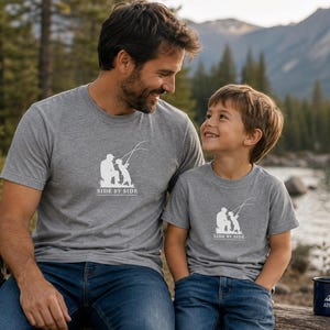 May include: Two gray t-shirts with a white graphic of a person and child fishing, with the text "SIDE BY SIDE". The shirts are worn by a man and a child, both wearing blue jeans. A blue mug with the word "ADVENTURE" is also visible.