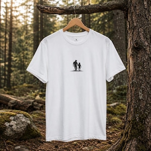 Può includere: T-shirt bianca con girocollo e una piccola silhouette nera di un genitore e un bambino che camminano insieme. La maglietta è appesa a una gruccia di legno su uno sfondo sfocato di foresta.