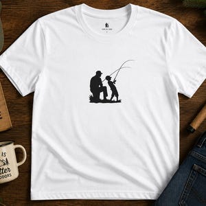 Può includere: T-shirt bianca con una silhouette nera di una persona e un bambino che pescano. La maglietta è circondata da accessori a tema outdoor, tra cui un berretto verde con la scritta "Adventure Awaits", un cartello di legno e una tazza con la scritta "Life is Better Outdoors".