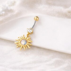 Könnte beinhalten: Ein goldfarbener Bauchnabelpiercing mit einem Sonnenanhänger. Die Sonne hat spitze Strahlen und eine schillernde Mitte. Der obere Ball und der hängende Edelstein sind klar. Körperschmuck.