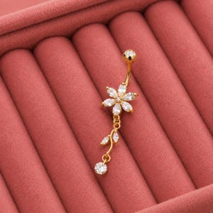 Könnte beinhalten: Goldfarbener Bauchnabelring mit floralem Design. Der Ring hat eine gebogene Langhantel mit einem kleinen, runden Oberteil und einem hängenden Blumenanhänger. Die Blume besteht aus klaren, funkelnden Steinen, mit einem einzelnen hängenden Stein unten.