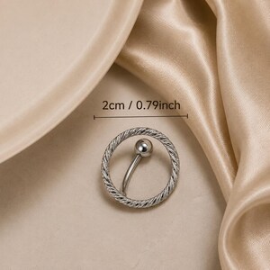 May include: A silver-toned belly button ring featuring a circular charm with a twisted rope texture and a curved barbell with a smooth ball end. The circular charm measures 0.79 inches in diameter, as indicated by the text 2cm / 0.79inch.