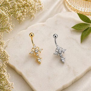 May include: Two belly button rings, one gold-toned and one silver-toned, each featuring a cross design adorned with clear gemstones. The jewelry is displayed on a white surface, with floral accents in the background.