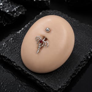 May include: A silver belly button ring with a red heart-shaped gem and a dangling, pointed design. The ring is displayed on a light-colored, oval-shaped surface, with a small silver stud above it. The background is a dark, textured surface.