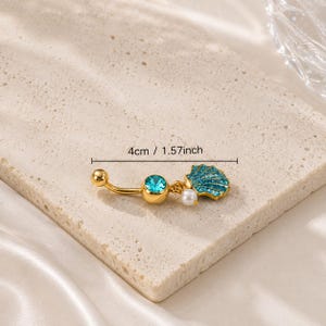 Puede incluir: Un piercing para el ombligo de color dorado con un diseño de barra curva. Cuenta con una gema turquesa, una pequeña perla blanca y un dije de concha azul. El anillo mide 4 cm de largo.