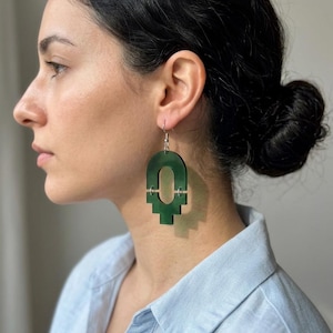 Könnte beinhalten: Geometrische grüne Acryl-Ohrringe mit silberfarbenen Haken. Die Ohrringe haben eine einzigartige, abstrakte Form mit einem zentralen Bogen und einem kreuzartigen Design am unteren Ende. Die Ohrringe sind tiefgrün.