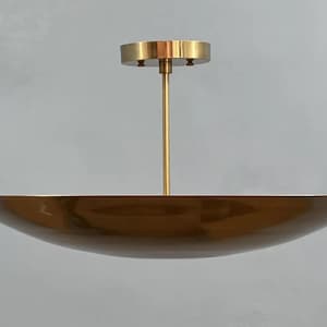 Op de afbeelding: Een moderne plafondlamp in goudtinten. De lamp heeft een grote, ondiepe komvormige kap die aan een centrale stang en een cirkelvormige plafondplaat hangt. Het ontwerp is minimalistisch en elegant, geschikt voor diverse interieurstijlen.