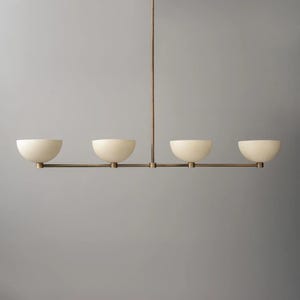 Può includere: Un lampadario moderno e lineare con quattro paralumi a forma di ciotola color crema. I paralumi sono sospesi a un telaio in metallo color bronzo. L'apparecchio è progettato per l'illuminazione dall'alto e aggiunge un tocco di eleganza minimalista.