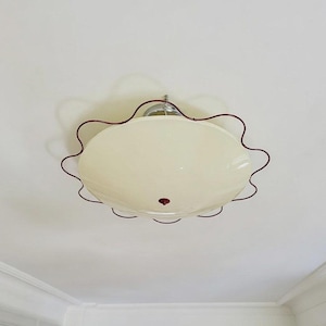 Puede incluir: Una lámpara de techo empotrada con una pantalla circular de cristal color blanco roto. La pantalla está rodeada por un marco fino de metal rojo oscuro con un borde ondulado y festoneado. Un pequeño remate circular rojo oscuro está centrado.