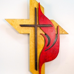 May include: Wooden wall art featuring a cross and a stylized red flame. The cross is dark brown, set against a light-colored wood background. The flame is a vibrant red, adding a striking contrast. The artwork is mounted on a white wall.