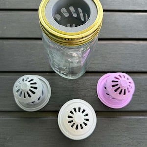 May include: A clear glass jar with a gold-colored lid and a gray filter. Three additional filters in gray, white, and lavender are also present. The filters have a circular design with multiple vent holes.