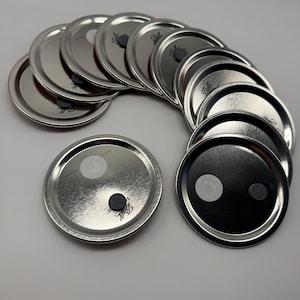 May include: A collection of circular silver-colored metal canning lids arranged in a fan shape. Each lid features a metallic finish, a small white circular sticker, and a dark gray circular button-like component with the Ball brand name printed.