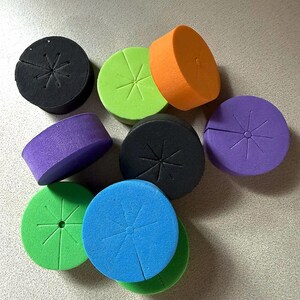 May include: A collection of colorful, round foam inserts in various colors, including black, green, orange, purple, and blue. Each insert has a star-shaped slit on the top, likely for hydroponic or gardening use.