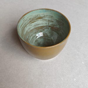 Può includere: Una ciotola in ceramica fatta a mano con una forma arrotondata. L'esterno è di un marrone caldo e terroso, mentre l'interno presenta una glassa strutturata, screziata di turchese e marrone. Il bordo della ciotola è leggermente più scuro.